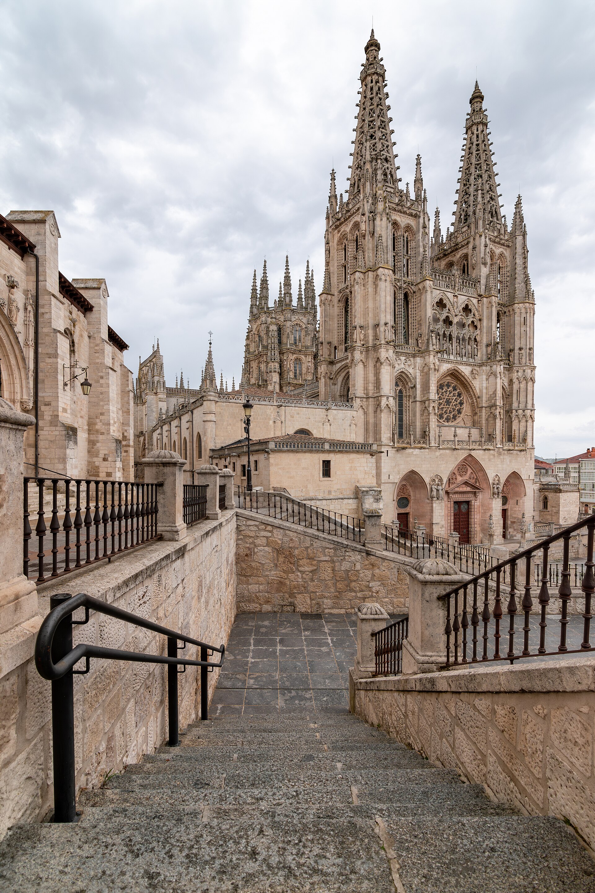 Escalinata hacia la catedral de Burgos: barandillas de hierro, dos agujas góticas y cielo cubierto.