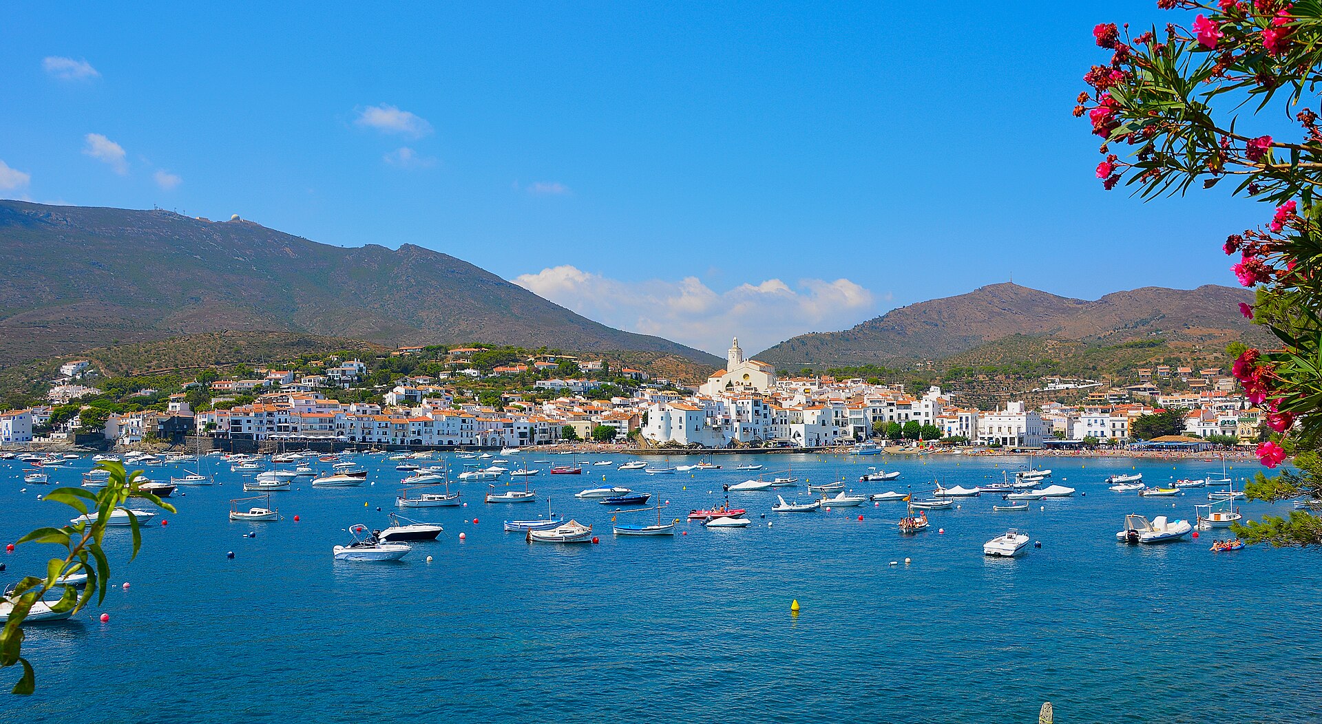 Bahía de Cadaqués: casco blanco, iglesia, barcas y adelfas en flor, montañas detrás.