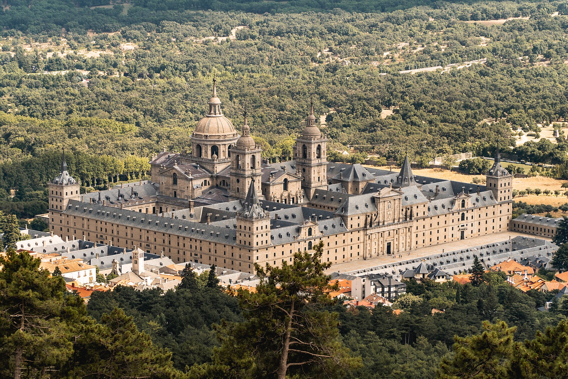 Monasterio de El Escorial visto desde arriba: planta en parrilla, cúpulas y bosque; tejados del pueblo delante.